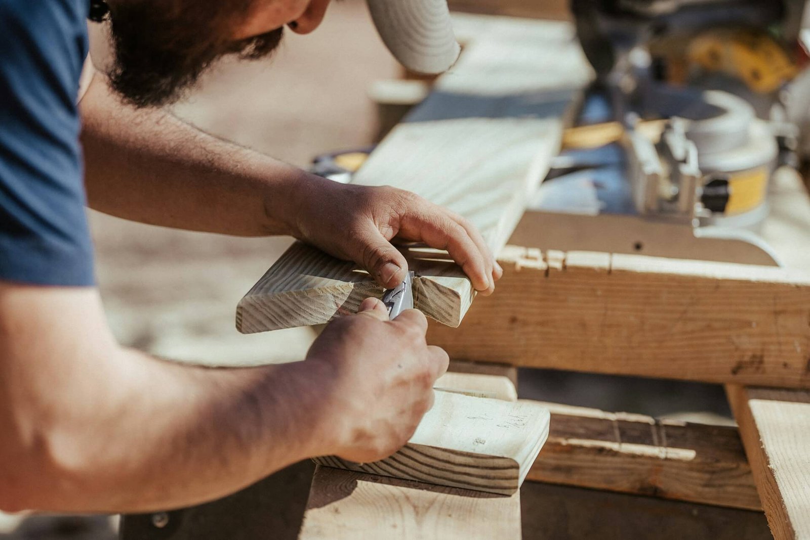 A person uses a pencil to mark a measurement on a wooden board, preparing to cut it on a workbench outdoors.